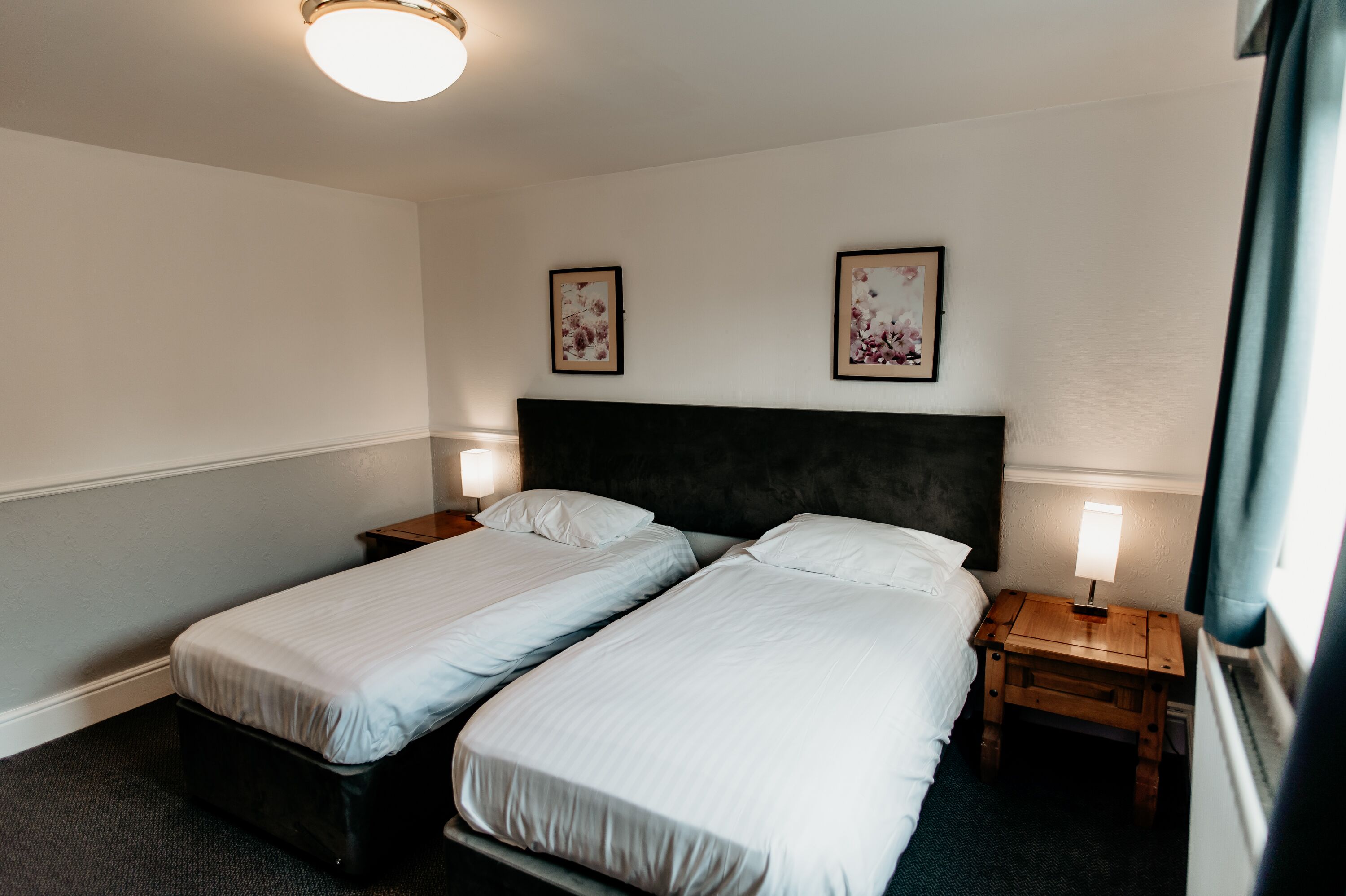 A hotel room with two single beds, a dark headboard, bedside lamps, and framed floral artwork on the wall.