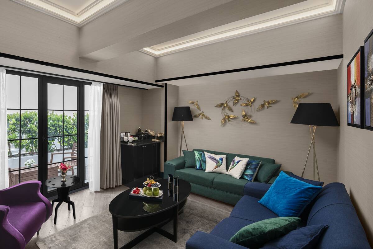 Una sala moderna con un sillón morado, un sofá azul y un sofá verde. Hay una mesa de centro negra con fruta y un jarrón. También se ve una lámpara de pie negra y un adorno dorado en la pared.