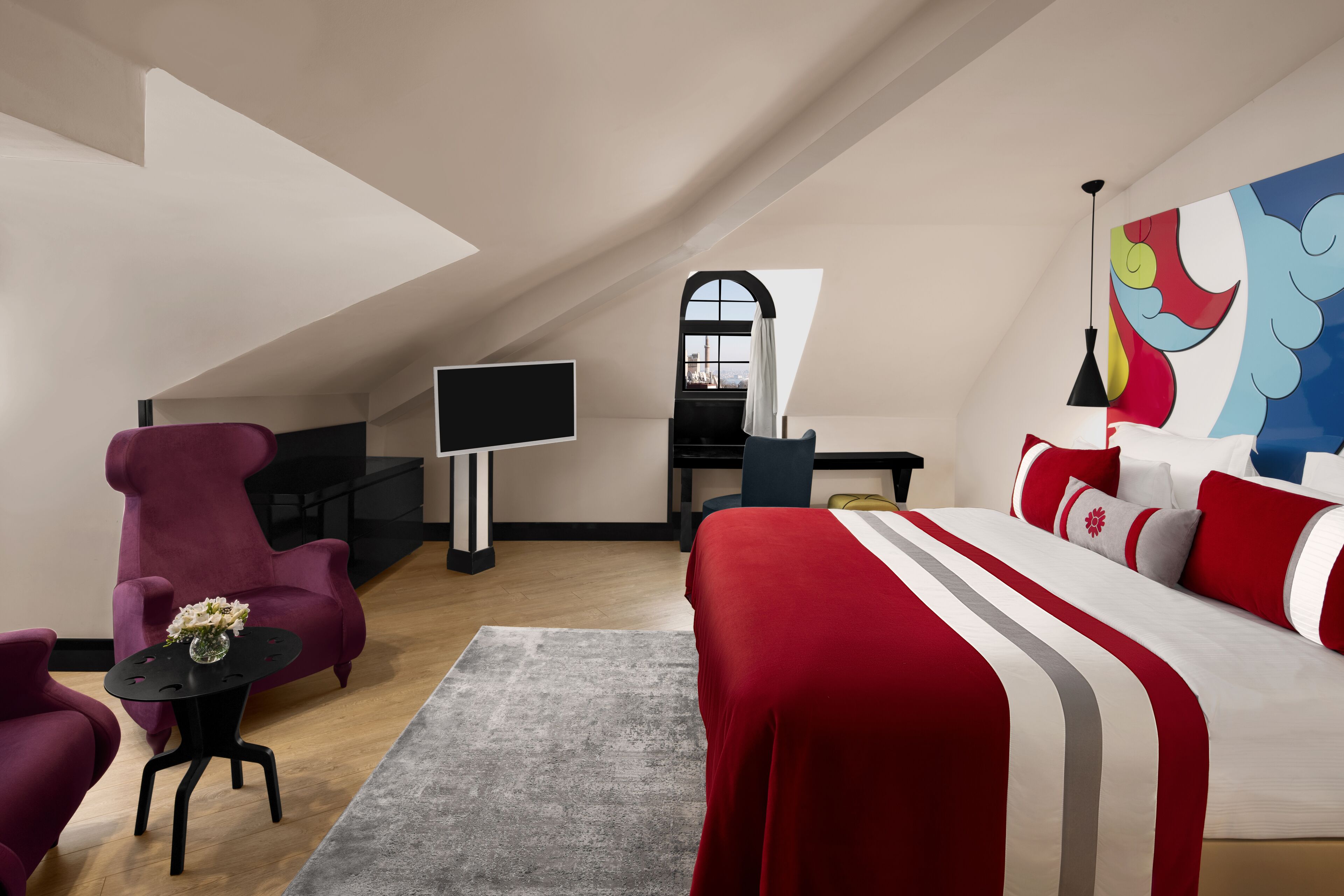 A modern bedroom with a sloped ceiling, a large bed with red and white bedding, a desk with a computer, a purple armchair, and a painting on the wall.