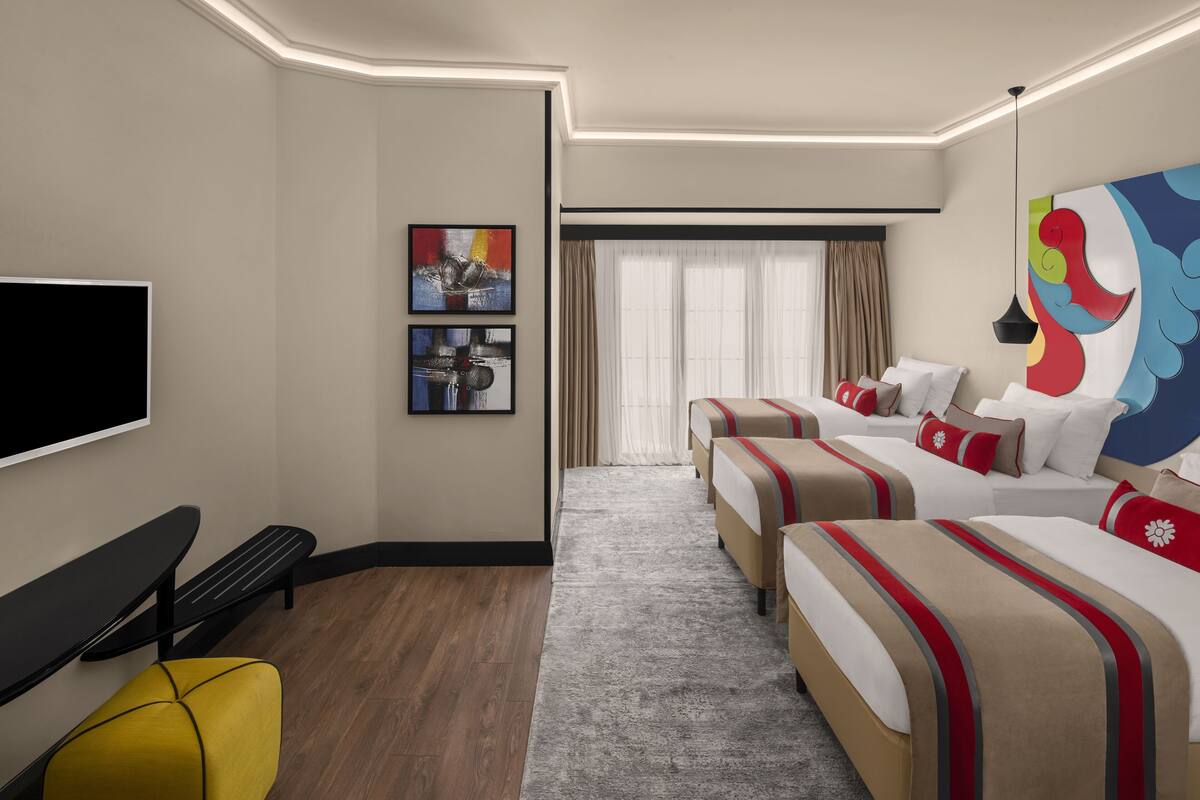 Habitación de hotel con televisión de pantalla plana, dos camas con ropa de cama roja y beige, un banco, una silla amarilla y obras de arte en las paredes.