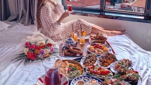 Iemand zit op een bed met een tafel vol eten en drinken, een raam met uitzicht op een stadsbeeld.