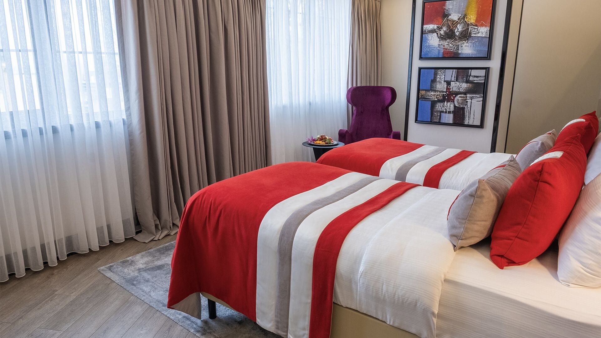 Habitació d'hotel amb dos llits, una cadira morada i una taula petita amb una cistella de fruita.