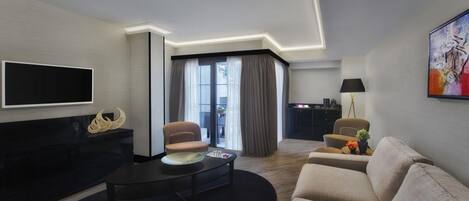 Una habitación de hotel moderna con una televisión de pantalla plana, un sofá, una mesa de centro y un área de comedor con una lámpara y un cuadro.