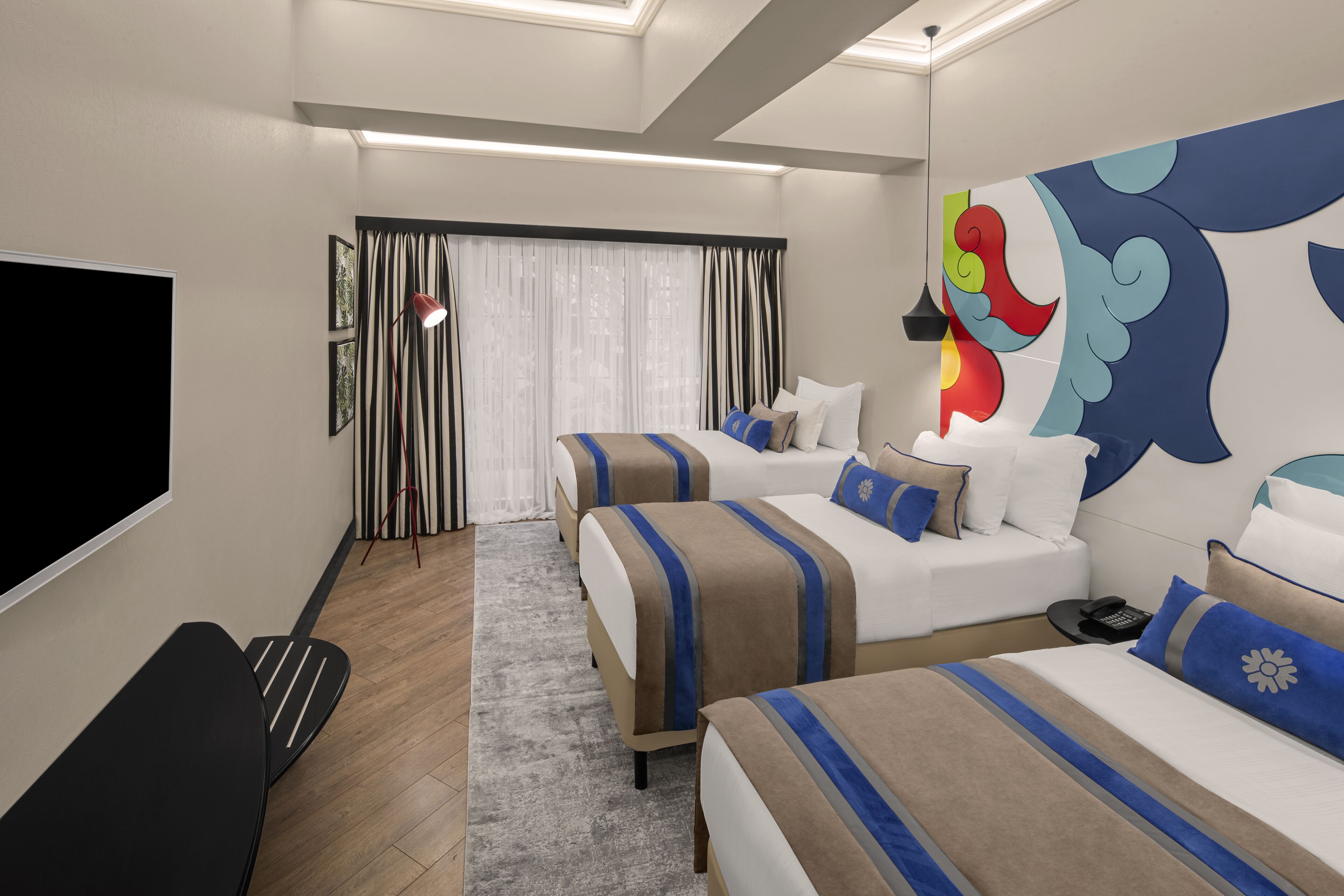 A hotel room with two beds, a flat-screen TV, a desk, and a large abstract wall mural.