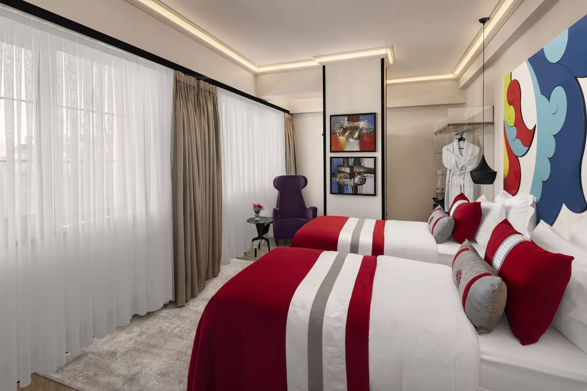 Habitación de hotel con dos camas, un ventanal con cortinas translúcidas, una mesita con un florero y un cuadro abstracto de colores en la pared.