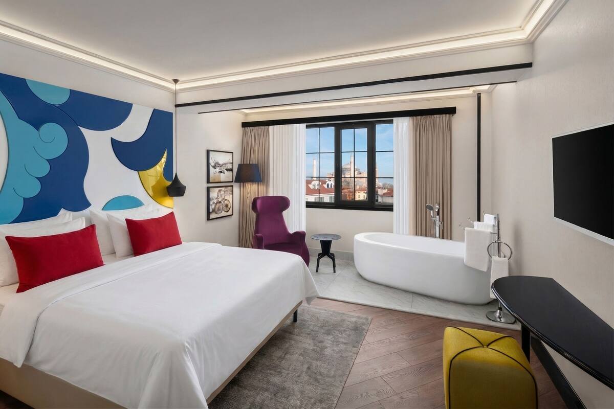 Habitación de hotel con cama, bañera, una mesita, una silla y televisión.