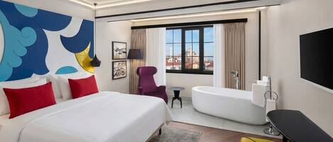 Habitación de hotel con cama, bañera, una mesita, una silla y televisión.