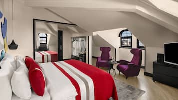A modern hotel room with a large bed, a desk, two purple armchairs, and a TV.