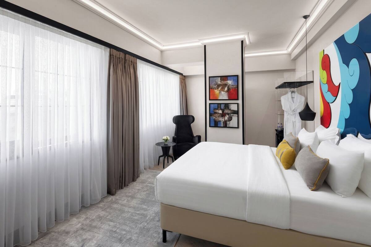 Una habitación de hotel moderna con una cama grande, una mesita con un florero, una silla y un cuadro abstracto de gran tamaño en la pared.