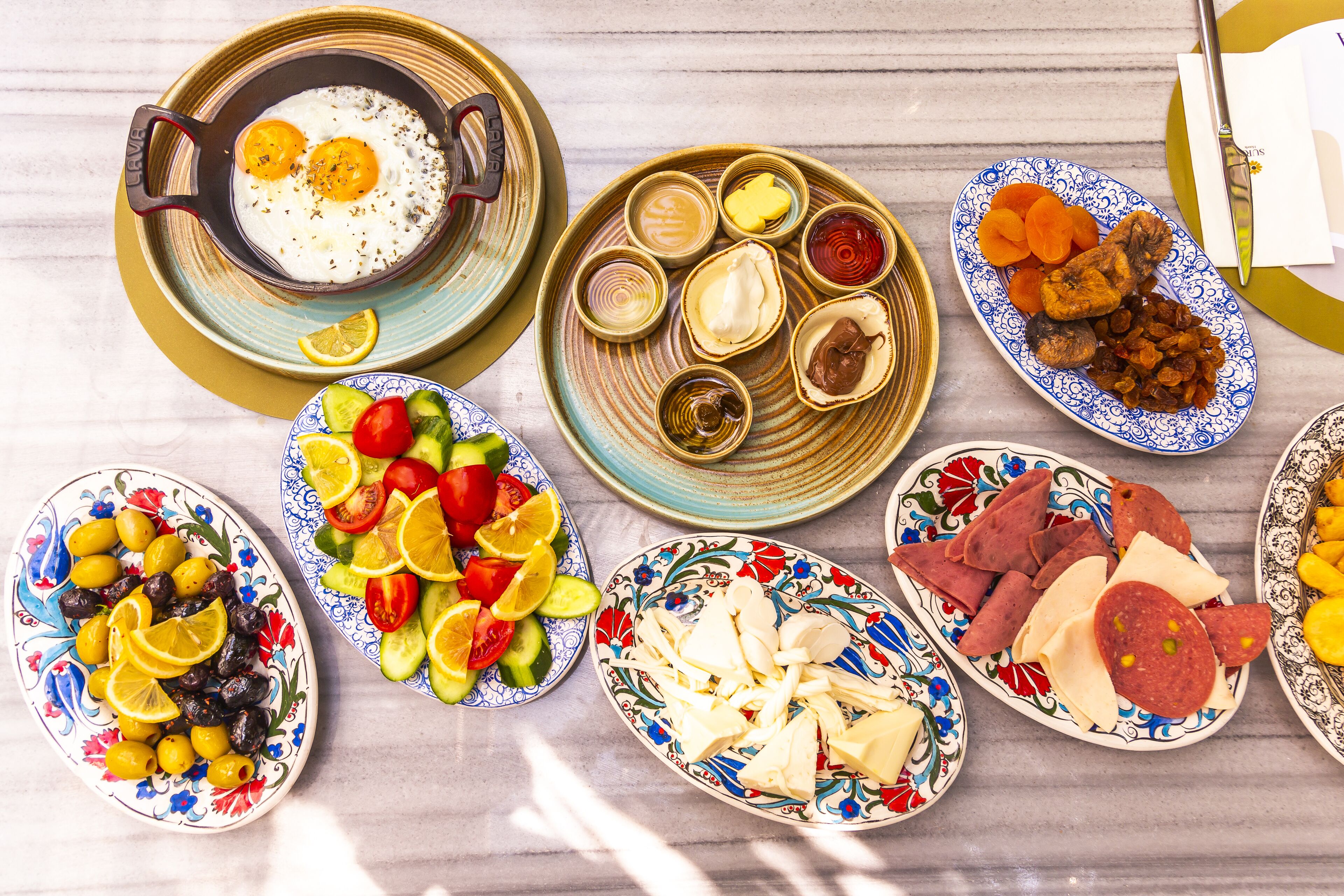 A variety of dishes on a table, including eggs, olives, tomatoes, cucumbers, cheese, and meats.