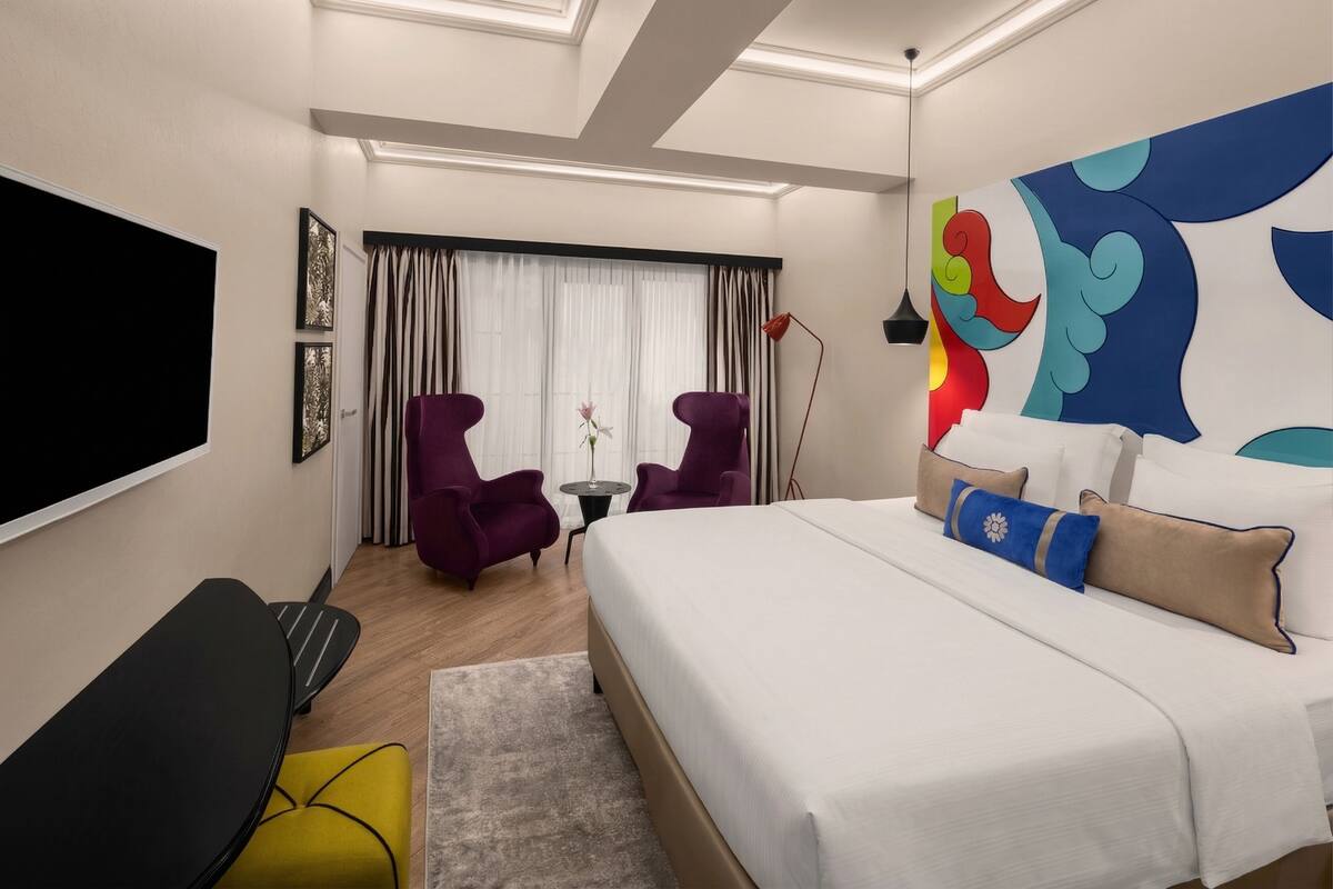 Una habitación de hotel moderna con una cama grande, dos sillones, una mesita, televisor y un mural colorido en la pared.
