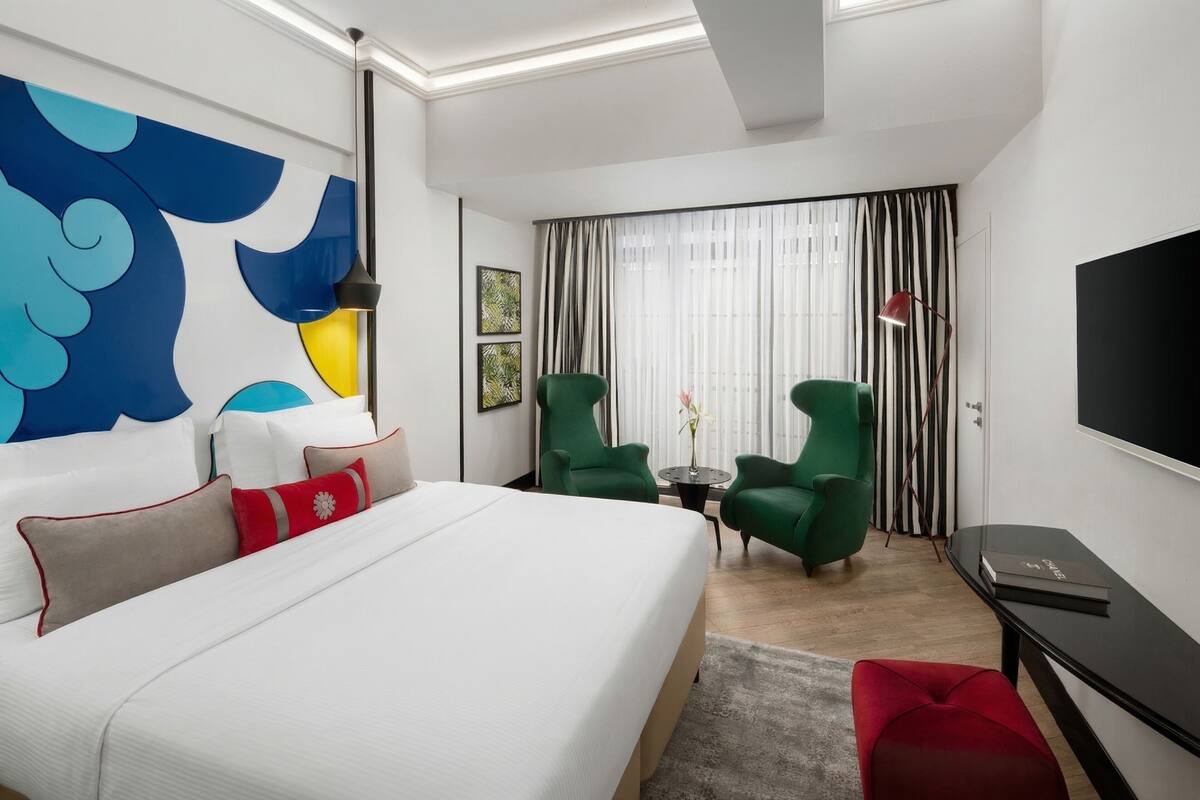 Una habitación de hotel moderna con una cama grande, sillones verdes, una mesita pequeña, un televisor y un cuadro abstracto de gran tamaño en la pared.