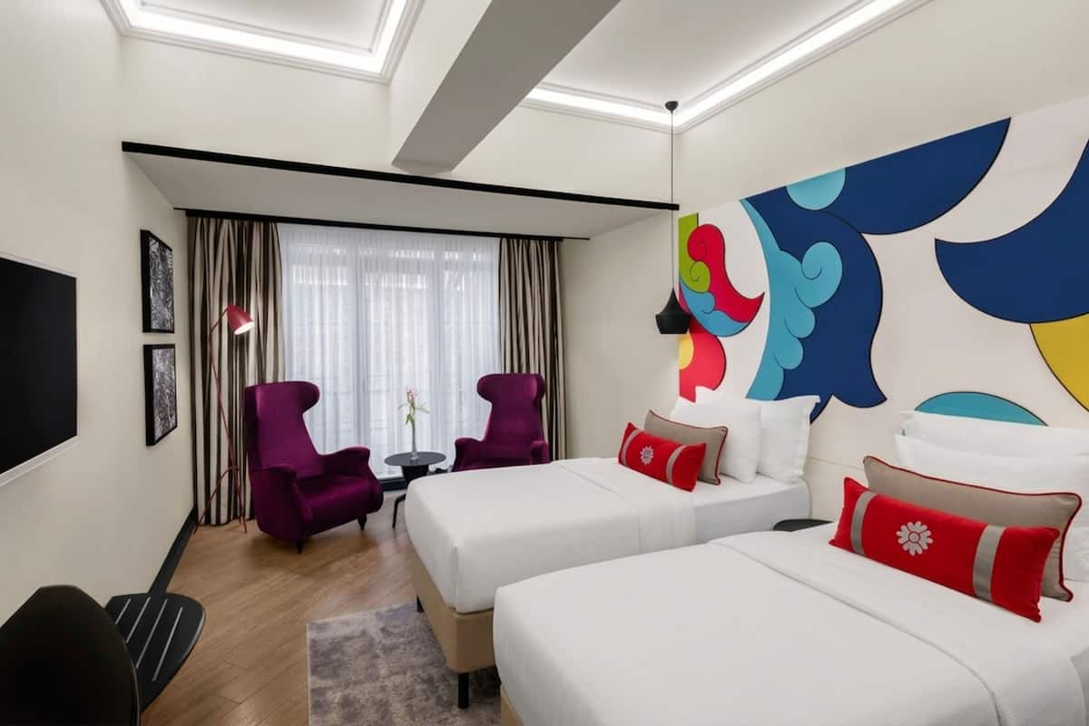 Habitación de hotel con dos camas, un televisor, un sillón púrpura, una mesita con un florero y un cuadro abstracto grande en la pared.