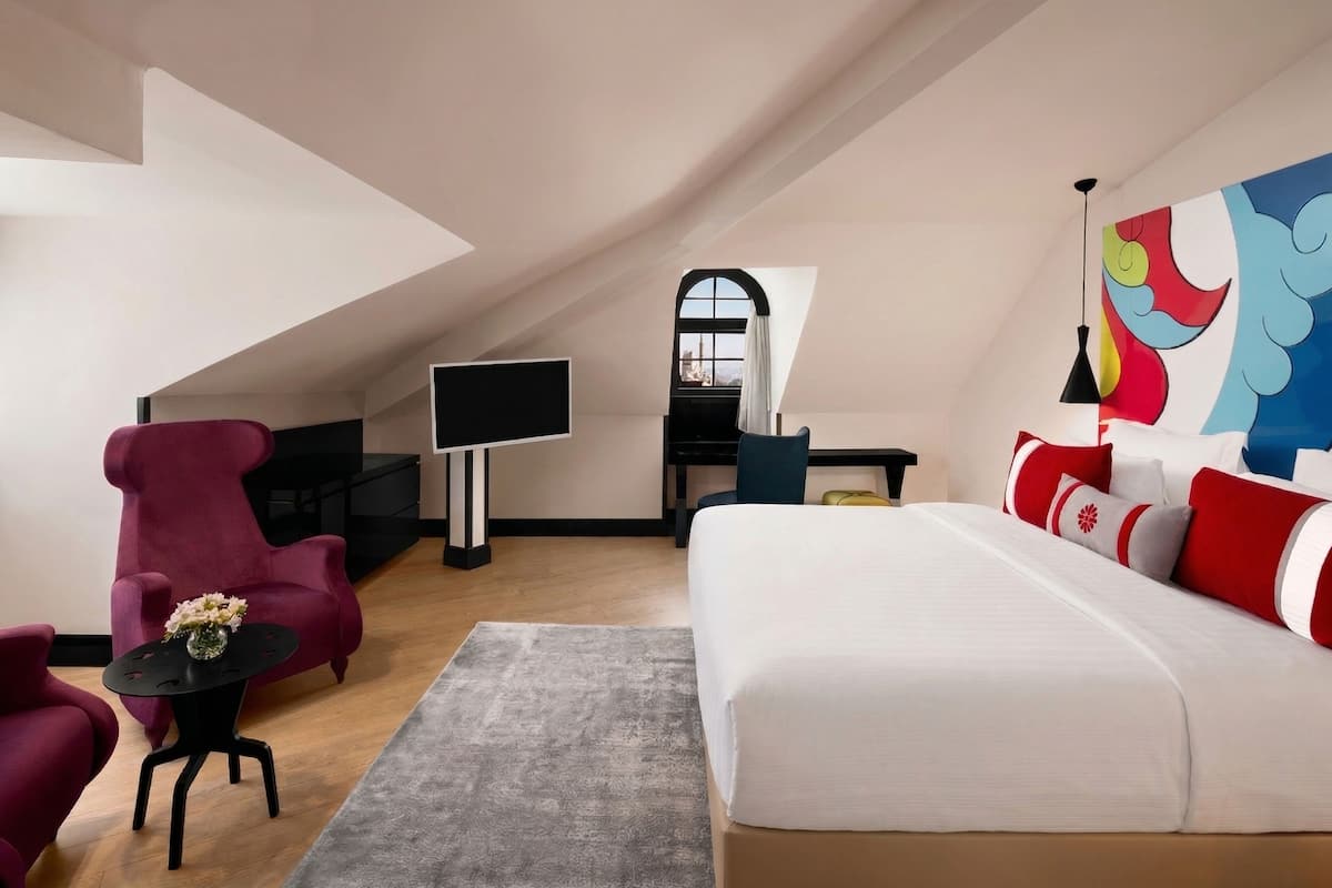 Habitación de hotel moderna con una cama grande, un escritorio con televisor de pantalla plana, un sillón rojo y un cuadro abstracto y colorido en la pared.