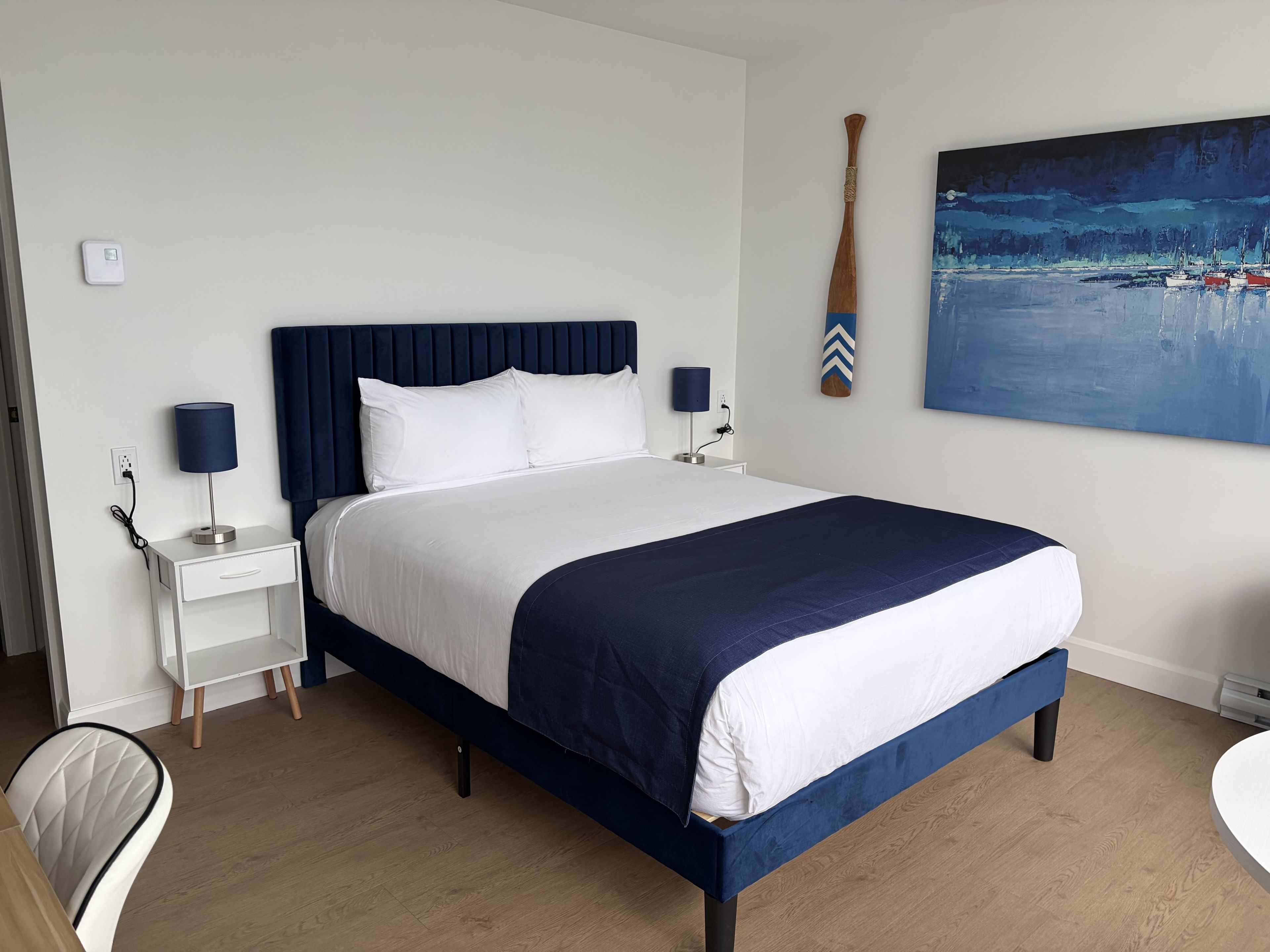 A neatly made bed with a dark headboard, white bedding, and a blue accent. There are two bedside tables with lamps, a chair, and a painting on the wall.