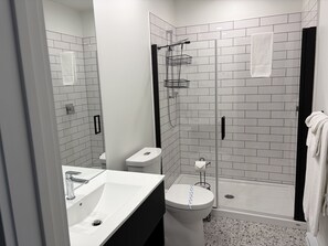 Moderni kylpyhuone, jossa on lasiseinällä varustettu suihku, valkoisilla laatoilla päällystetyt seinät, valkoinen pesuallas ja wc-istuin.