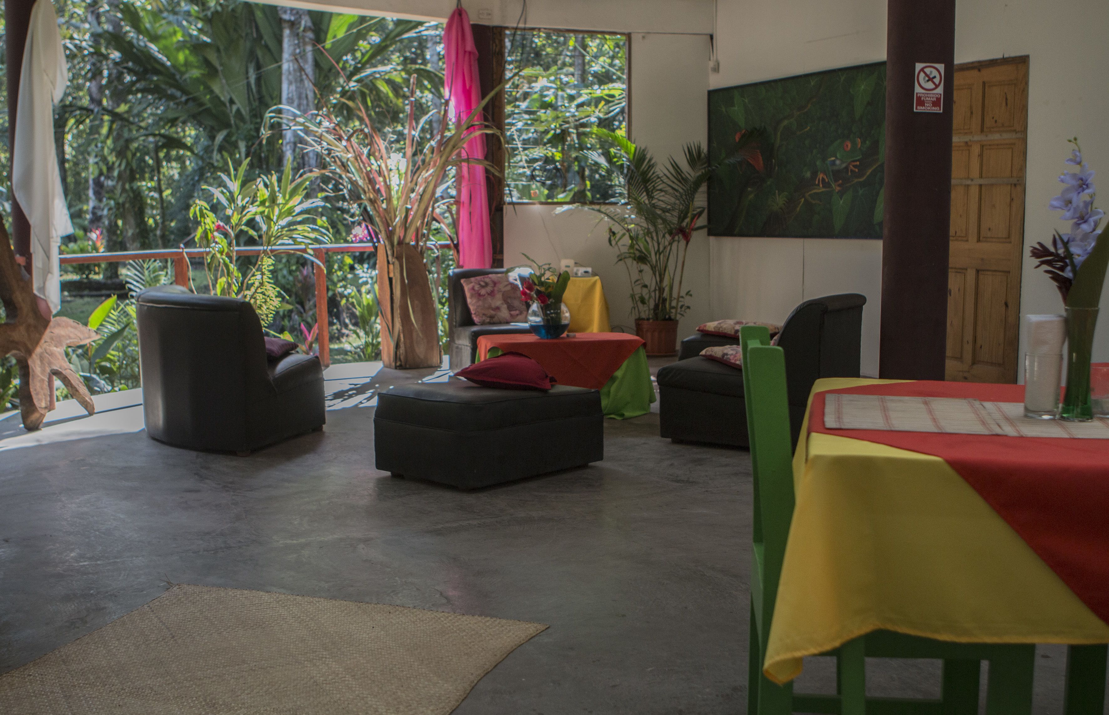 A room with a concrete floor, a table with a red and yellow tablecloth, black and green chairs, and a large window with a view of greenery.