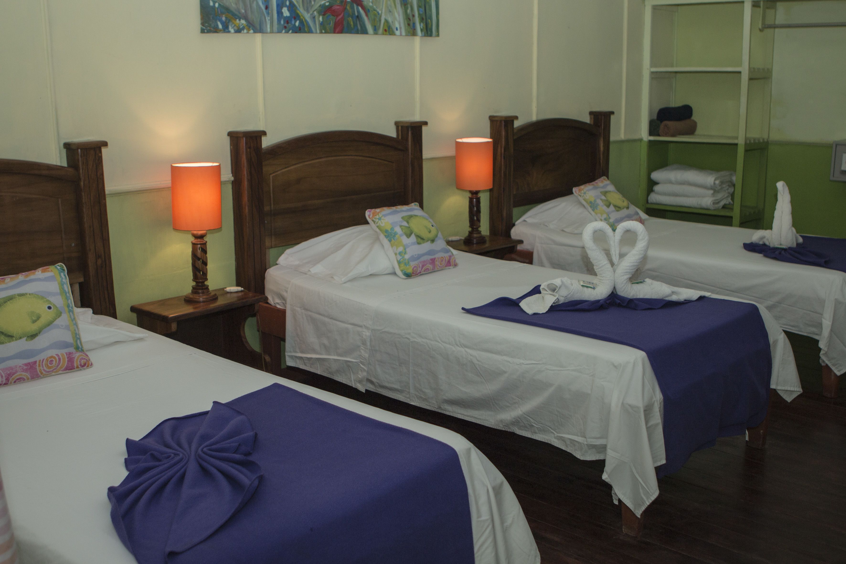 Habitación de hotel con dos camas, cada una con cabecero de madera, ropa blanca de cama y una manta púrpura en una de ellas. Hay dos lámparas de noche con pantallas naranjas y una pared verde con un cuadro.