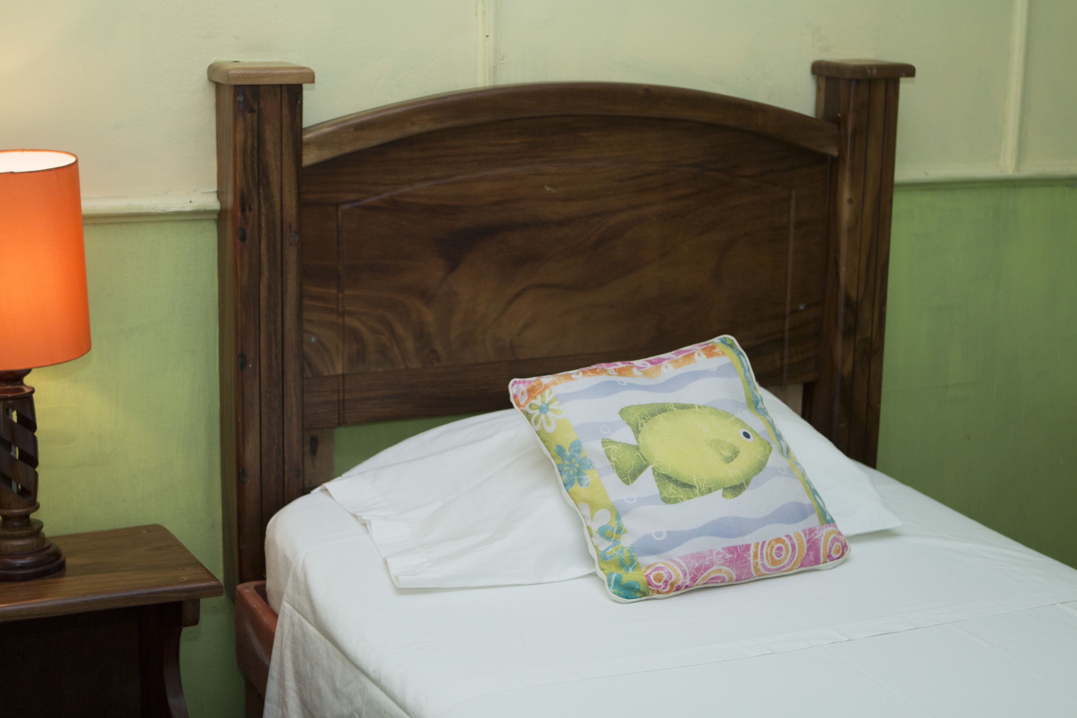 Un lit avec un drap blanc, une tête de lit en bois et un coussin coloré orné d’un motif de poisson.