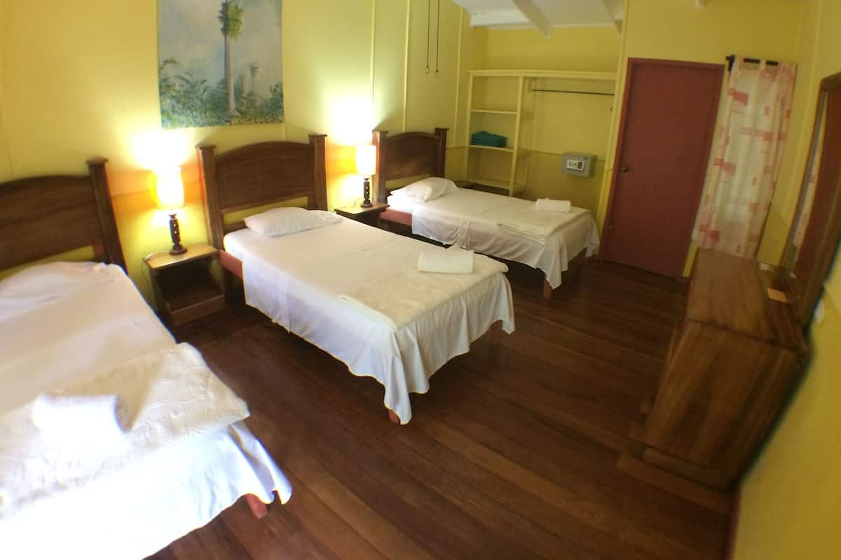 A hotel room with three single beds, wooden headboards, a wardrobe, a painting on the wall, and a bedside table with a lamp.