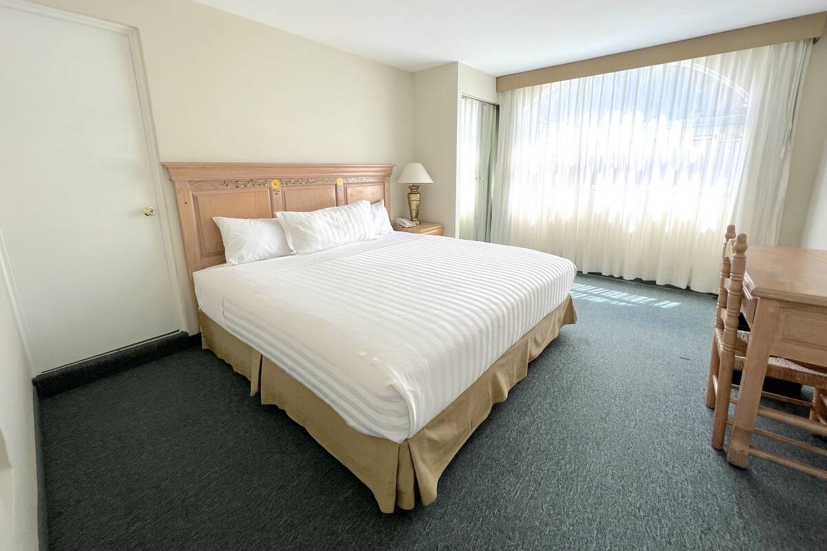 A hotel room with a large bed, a wooden headboard, a desk with a chair, and a window with curtains.