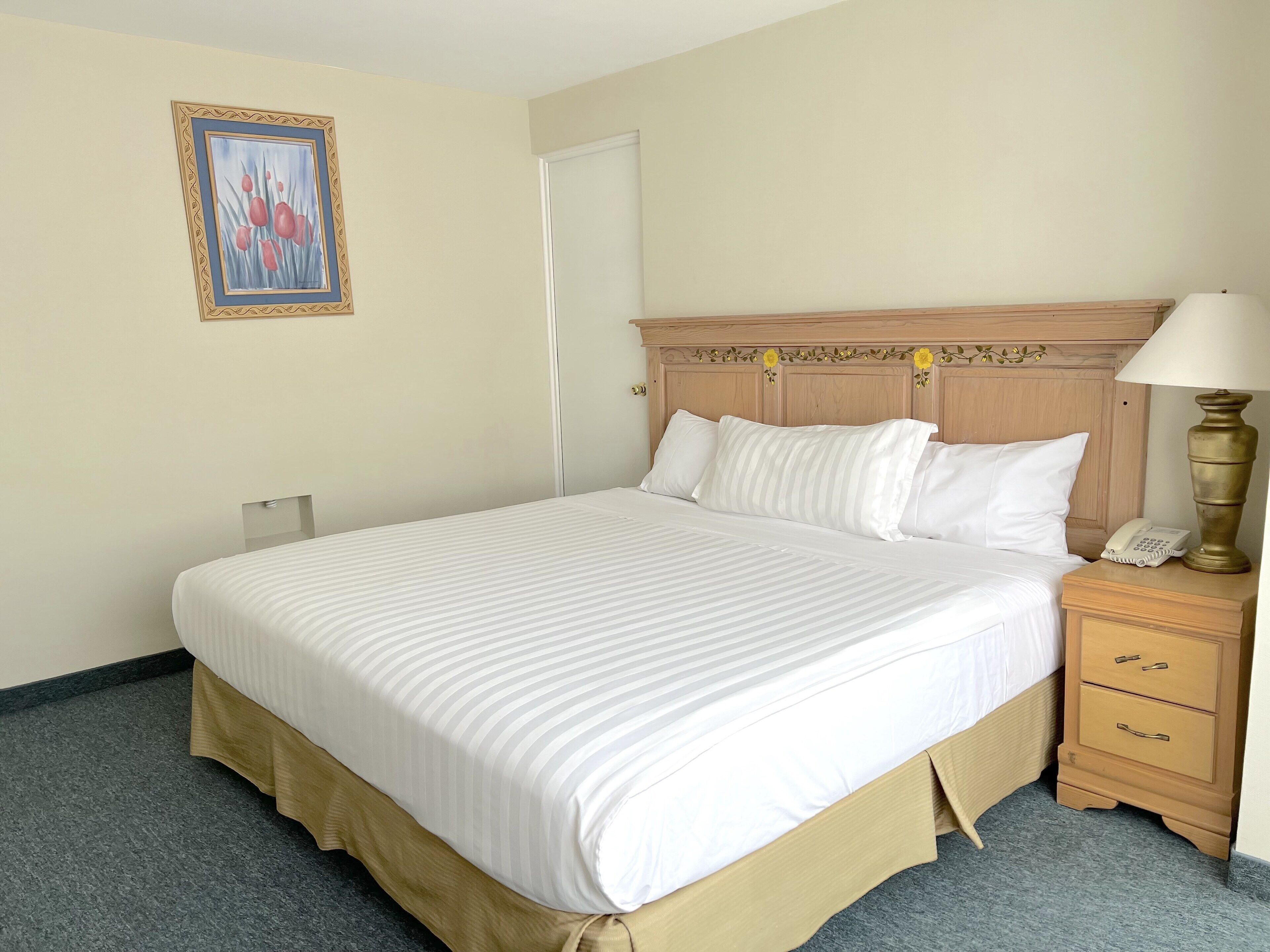 Quarto de hotel com uma cama grande, mesa de cabeceira de madeira com abajur, telefone e quadro na parede.