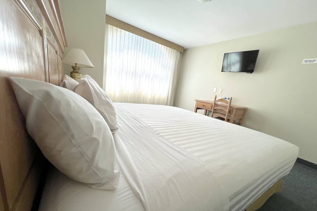 A hotel room with a bed, a desk, a chair, a lamp, and a TV on the wall.