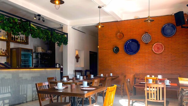 Um restaurante com mesas e cadeiras de madeira, parede de tijolos com pratos decorativos e um espaço de bar com prateleiras de garrafas.