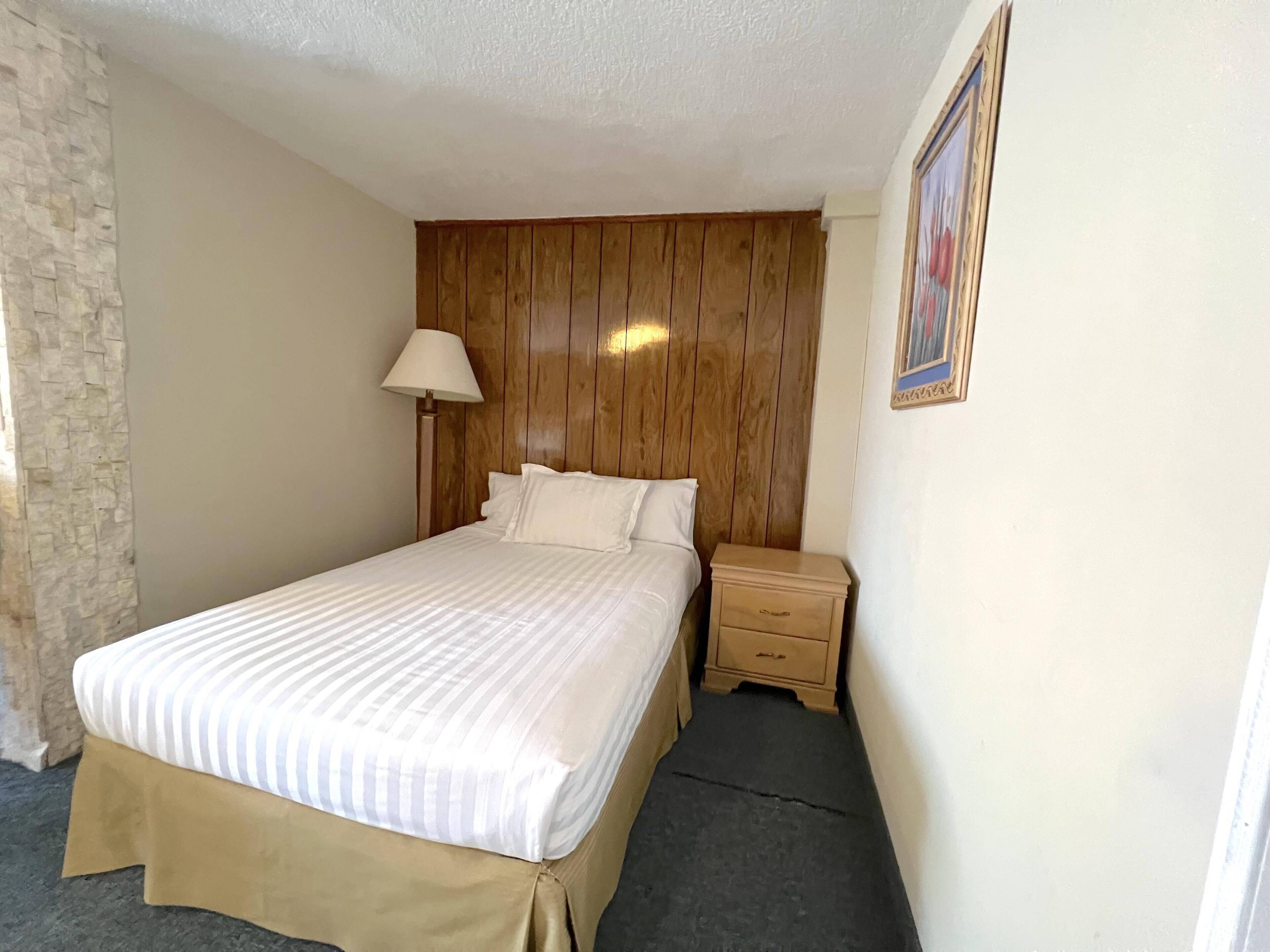 A neatly made bed with a striped bedspread, a wooden headboard, a bedside lamp, and a small wooden nightstand with a drawer.