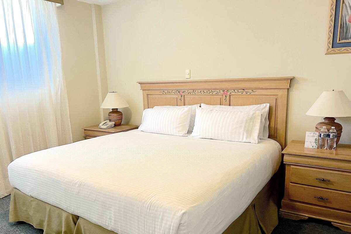 A neatly made bed with a wooden headboard, two bedside tables with lamps, a telephone, and a water bottle on one of the tables.