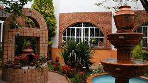 Pátio com parede de tijolos, uma fonte e plantas em vasos.