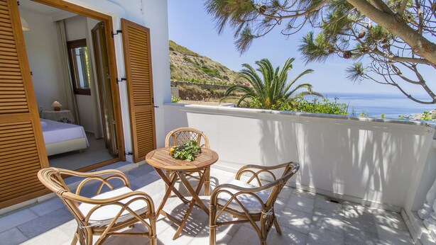 A balcony with wicker furniture, a small table with a fruit bowl, and a view of the sea and hills. - Petrino (Naxos)