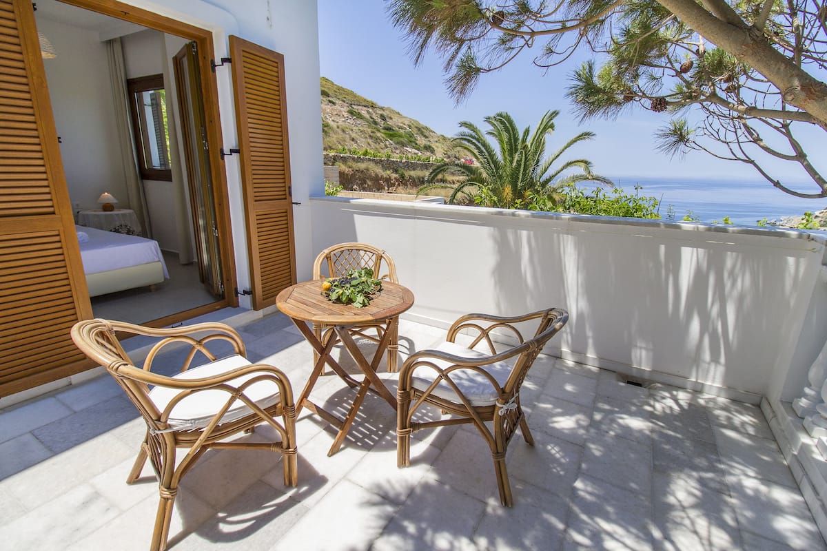 A balcony with wicker furniture, a small table with a fruit bowl, and a view of the sea and hills.