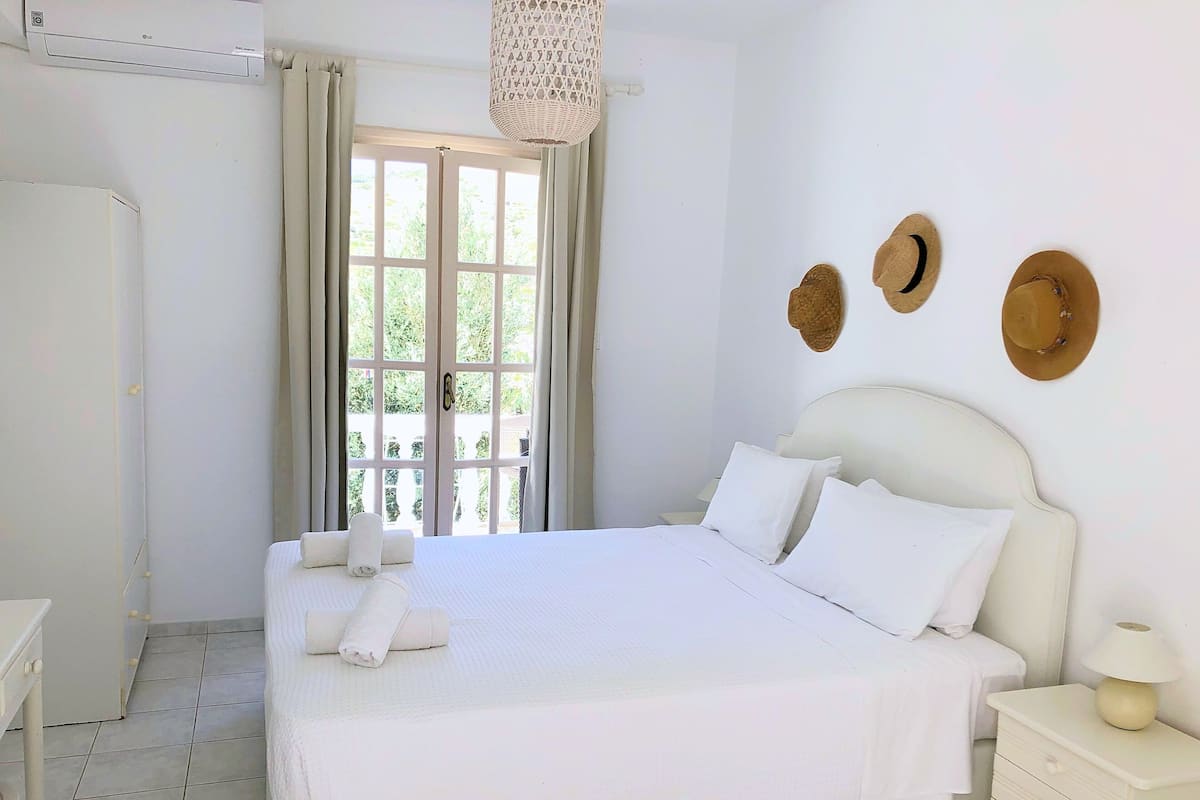 A neatly made bed with white linens, a bedside table with a lamp, and a view of greenery through a window with curtains.