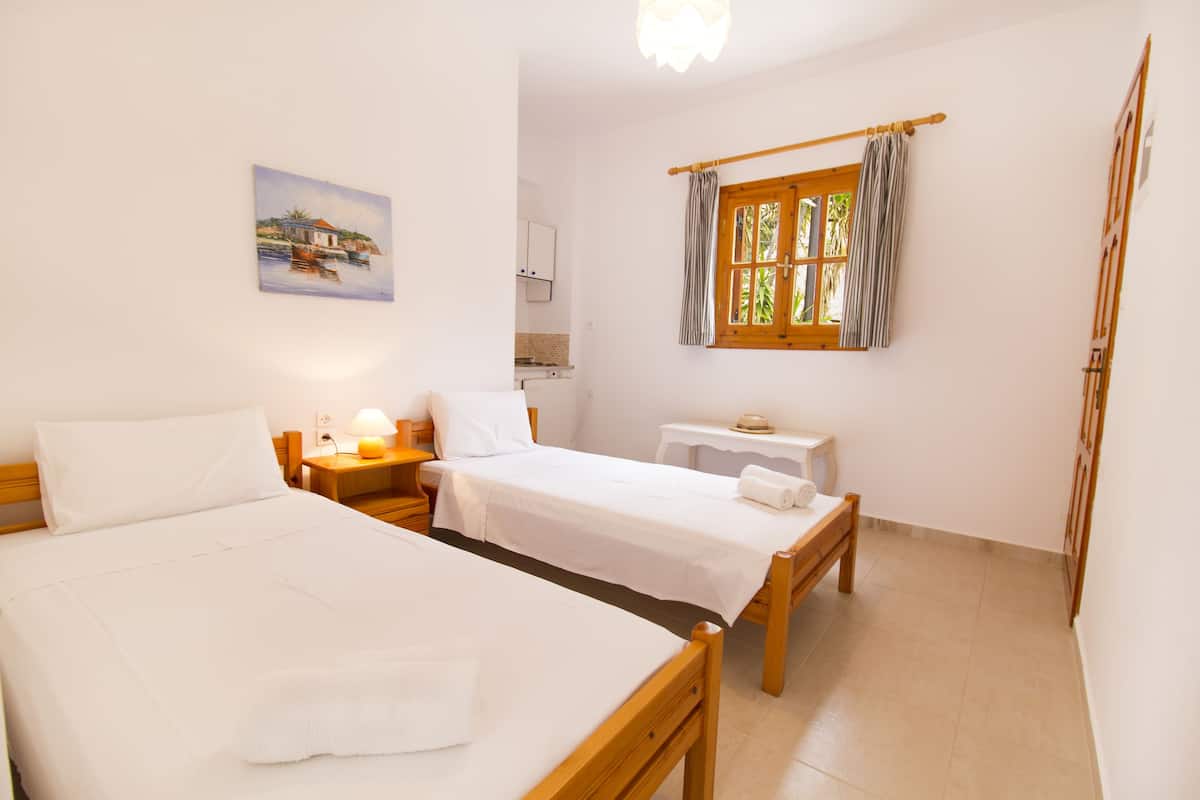 A hotel room with two single beds, a small table, a nightstand, a painting on the wall, and a window with curtains.