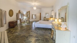 A spacious room with a stone floor, a large bed, a sitting area with a chair and table, and a mirror with a decorative frame. - Petrino (Naxos)