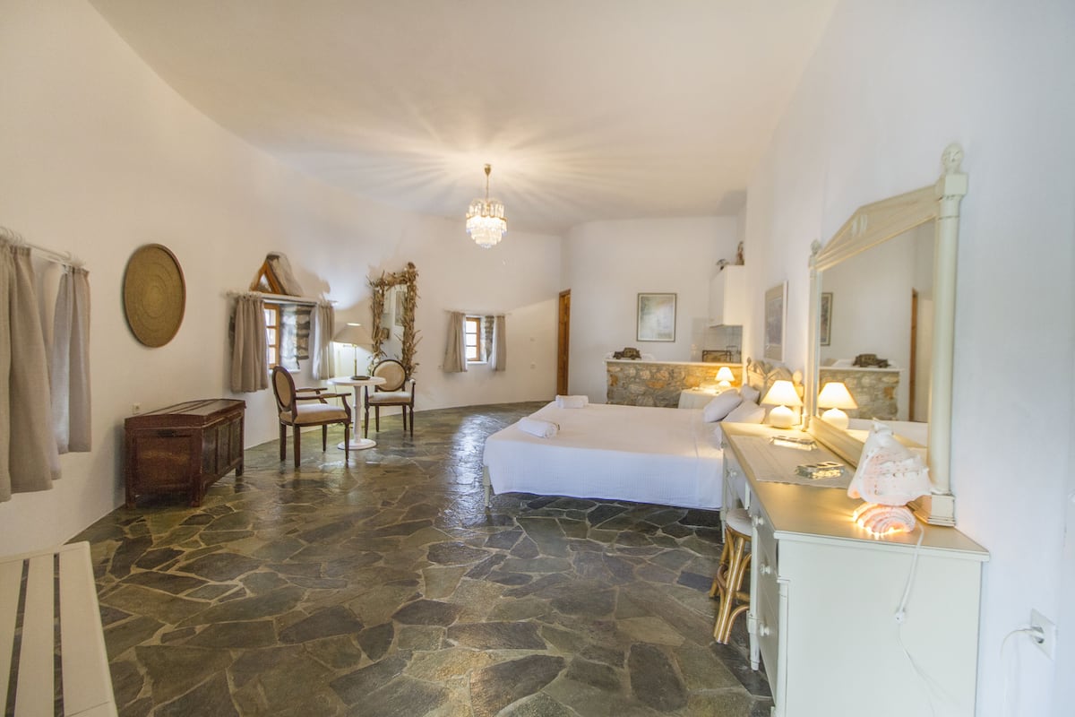 A spacious room with a stone floor, a large bed, a sitting area with a chair and table, and a mirror with a decorative frame.