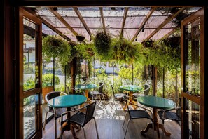 An indoor patio with glass tables and chairs, surrounded by hanging plants and a view of the street. - Hotel Boutique Britânico (Gramado)