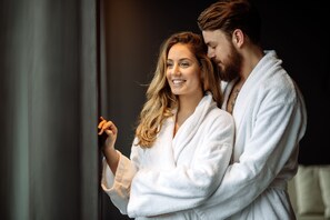 A couple in white bathrobes standing close together, possibly in a hotel room.
