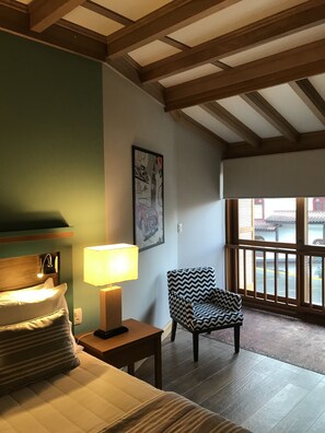A hotel room with a bed, a chair, a table, a lamp, a window, and a painting on the wall.