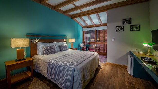 A bedroom with a bed, bedside tables, a desk, and a window with blinds. - Hotel Boutique Britânico (Gramado)