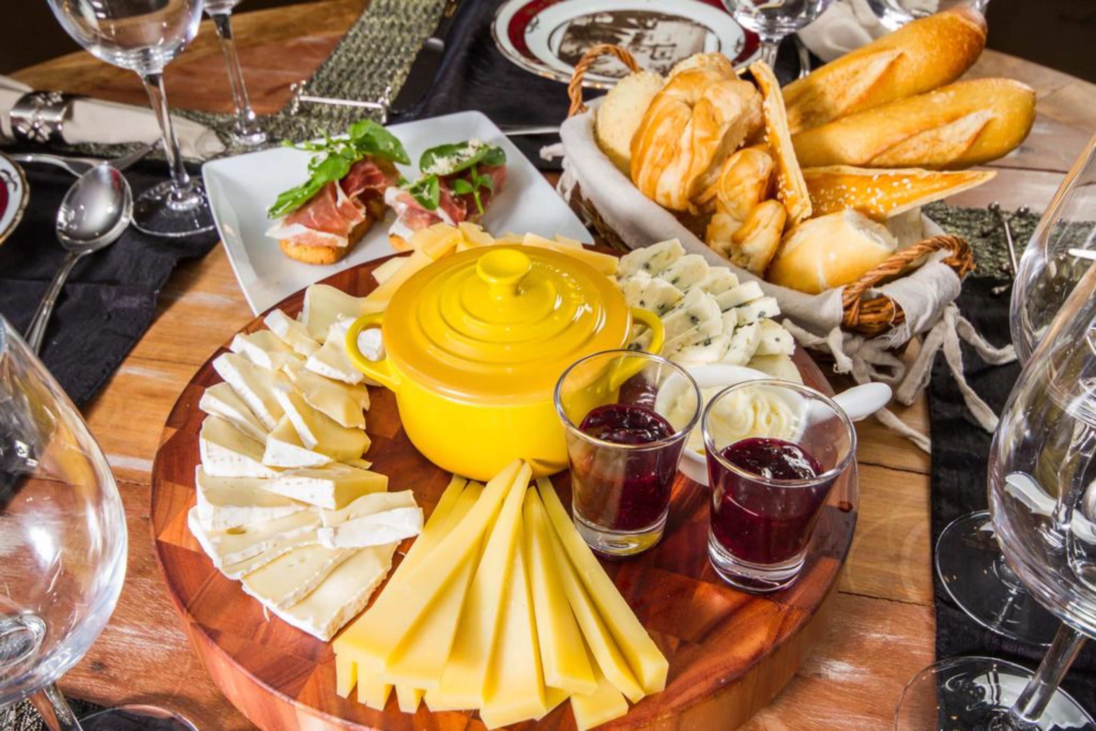 A charcuterie board with various cheeses, bread, and meats, accompanied by wine glasses and a yellow pot.