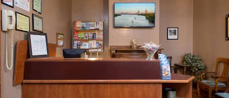 A reception desk with a wooden finish, a mounted television displaying a cityscape, and a bookshelf filled with books and magazines.
