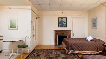 Um quarto com cama, uma cadeira, uma mesinha, lareira e um quadro na parede.