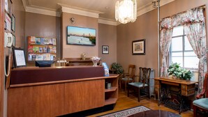 A reception area with a wooden desk, a chandelier, a large window with floral curtains, and a wall-mounted TV displaying a cityscape. - The Farrington Inn (Boston)