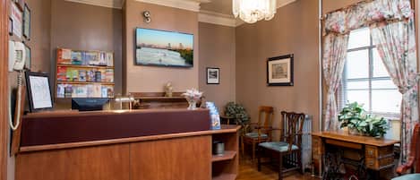 A reception area with a wooden desk, a chandelier, a large window with floral curtains, and a wall-mounted TV displaying a cityscape.