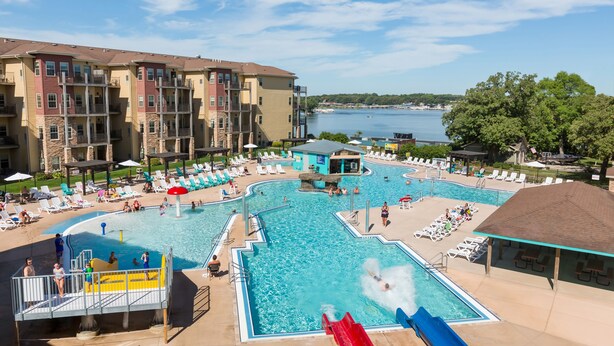 A swimming pool area with multiple water slides, lounge chairs, and a view of a lake. - Bridges Bay Resort (Arnolds Park)