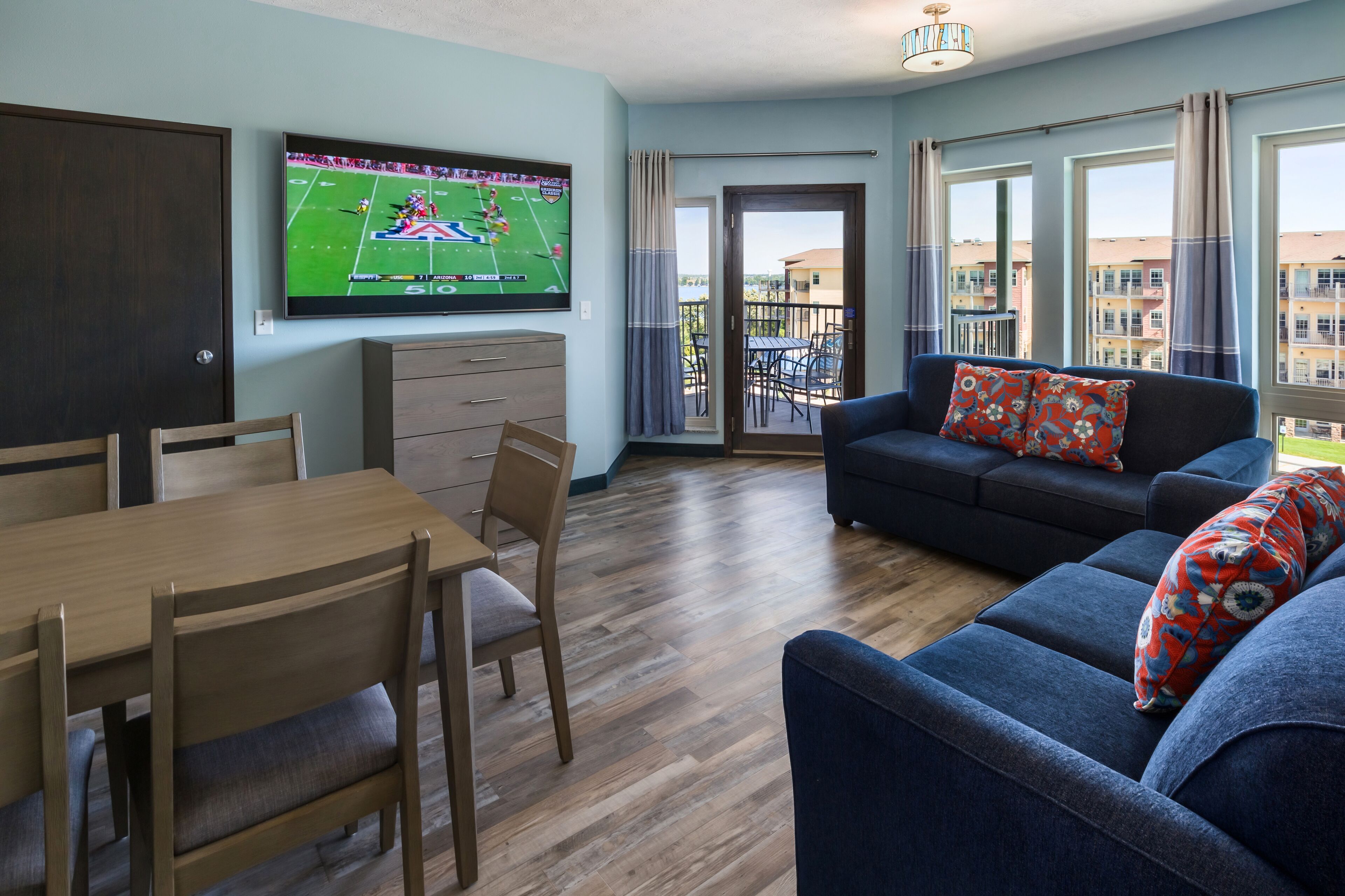 Un salon avec un téléviseur diffusant un match de football, un canapé, une table à manger avec des chaises et un balcon offrant une vue sur les immeubles.