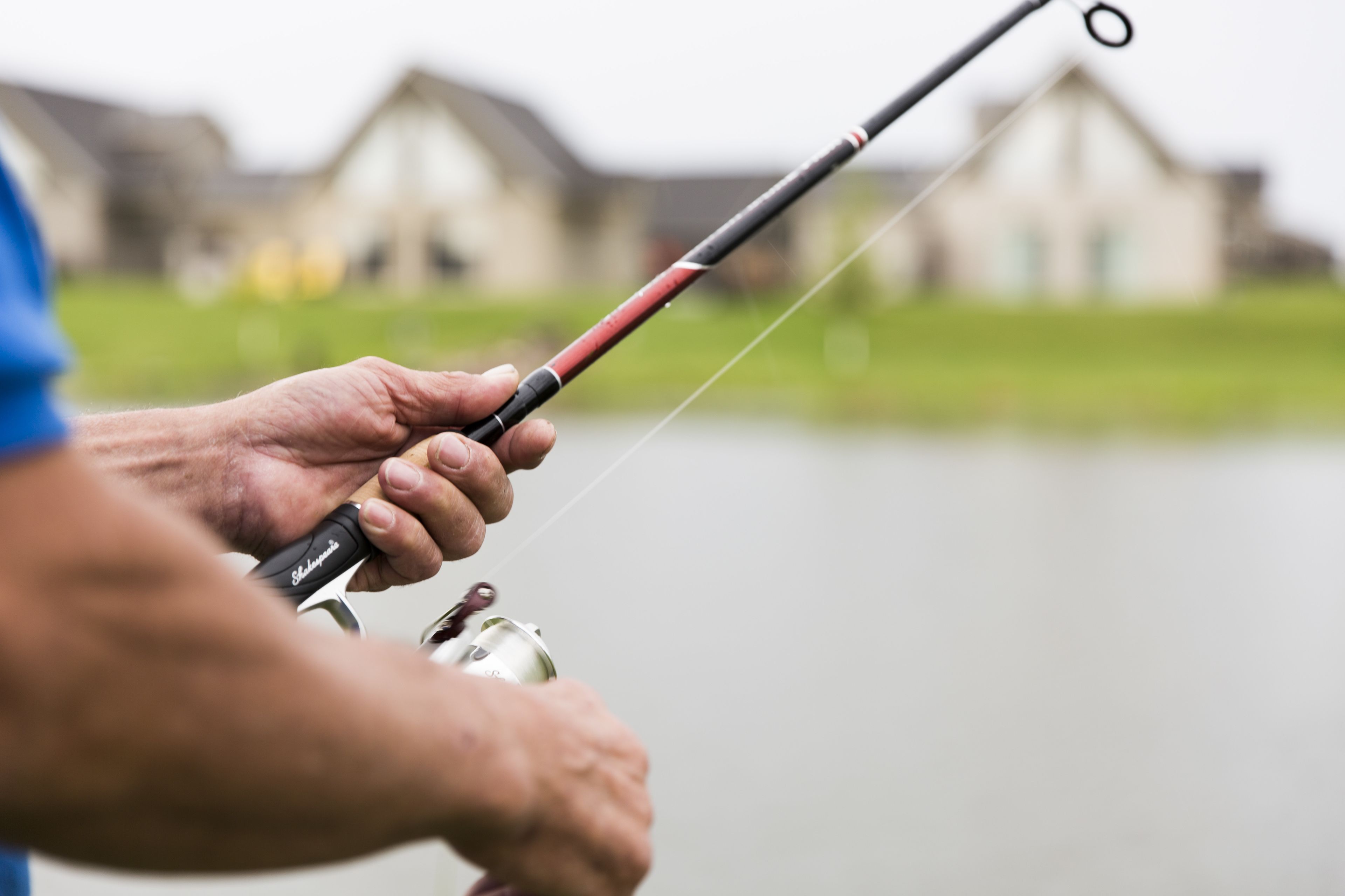 A person holding a fishing rod with a reel, standing by a body of water with houses in the background.