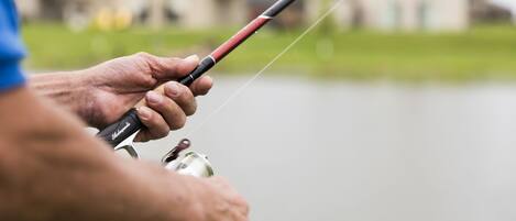 A person holding a fishing rod with a reel, standing by a body of water with houses in the background.